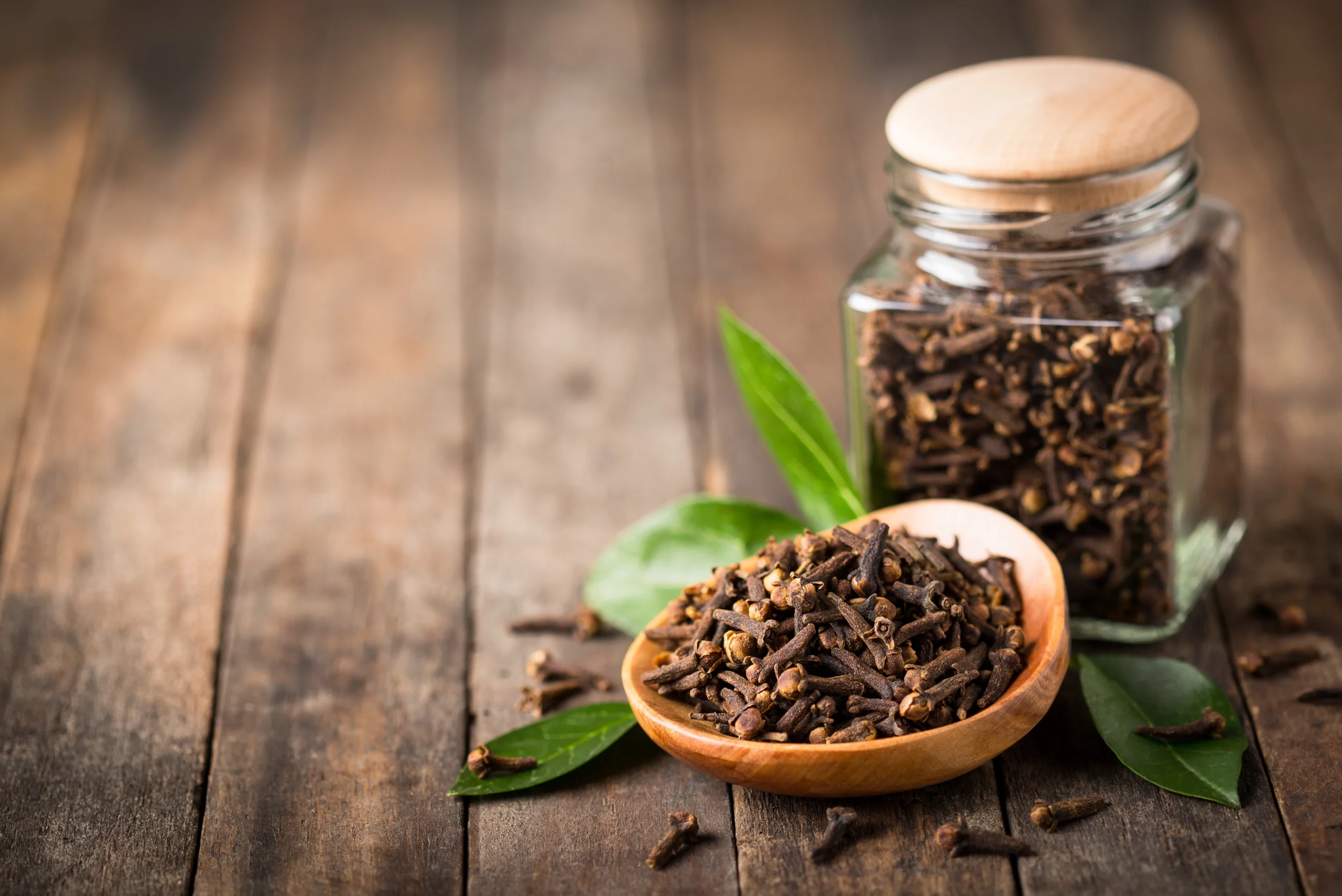 The image presents a rustic still life featuring cloves on a wooden spoon and jar